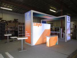 Custom Island Exhibit with Extensive Backlit Graphics, Layered Hanging Sign, Backlit Reception Counter, Mounted Edge-lit Counter with Drawer, Monitor Mounts, Painted Logos, (1) MOD-1456 Charging Table, and (2) MOD-1454 Charging Tables